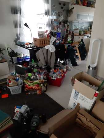 Unordentlicher Wohnraum mit vollgestelltem Tisch, Kartons, Verpackungen und Haushaltsgegenständen.